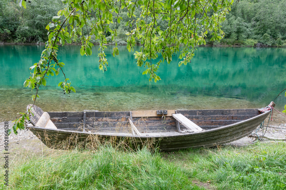 Old wooden boat on the river bank in Norway. Green grass and foliage, grey and turquoise water, mountains and cloudy sky on the background