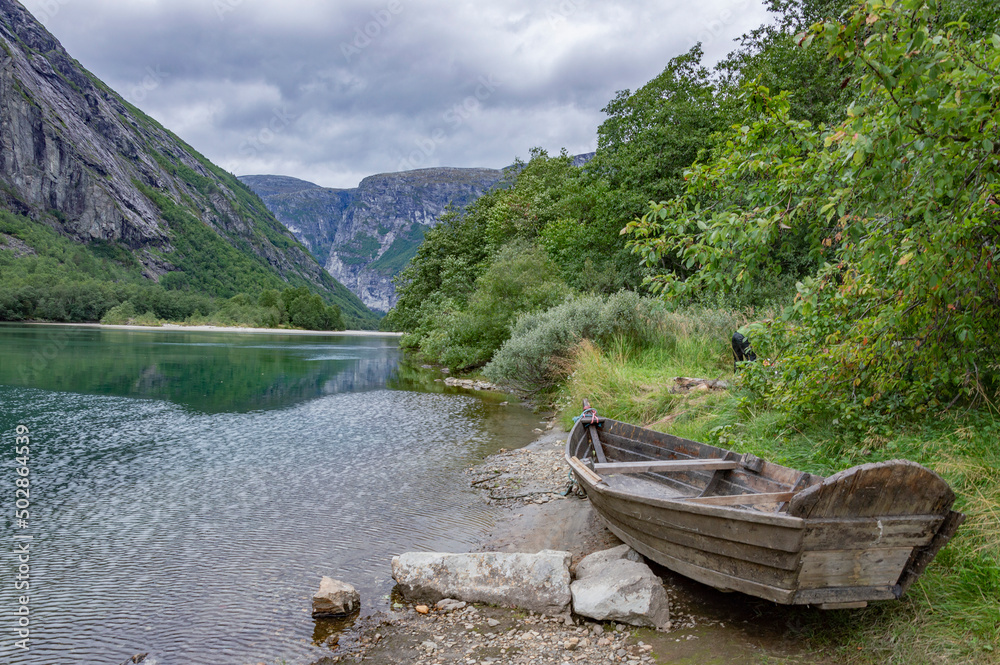 Fototapeta premium Old wooden boat on the river bank in Norway. Green grass and trees, clear water, mountains and cloudy sky on the background