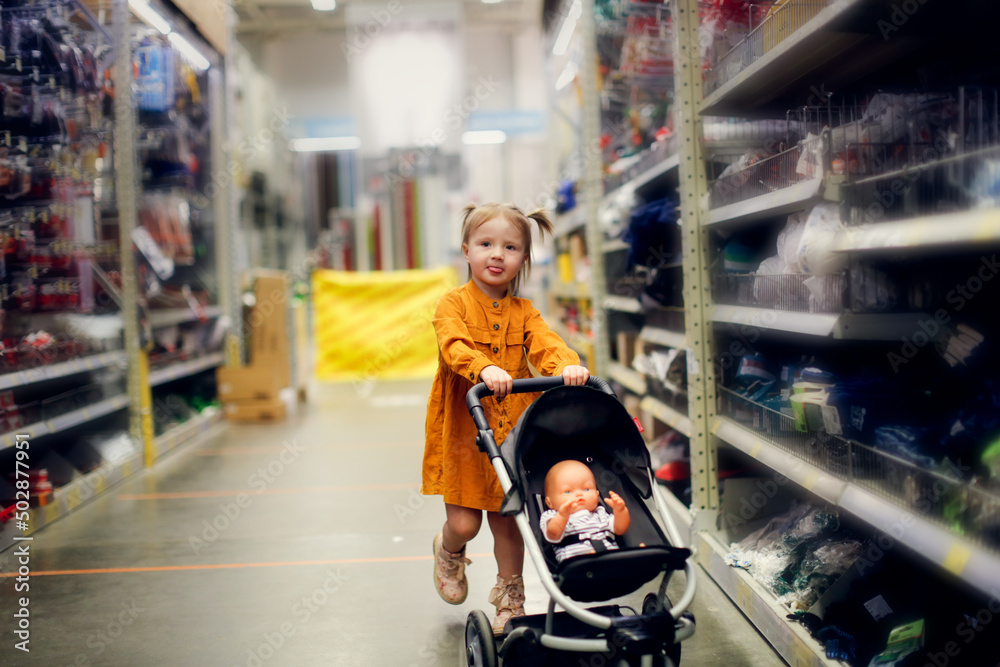 Cute baby girl in a mustard dress with doll in puppet stroller in a store, a child is playing in a hardware store