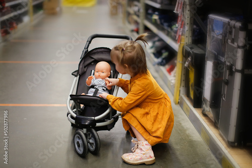 Obraz na plátně Cute baby girl in a mustard dress with doll in puppet stroller in a store, a chi