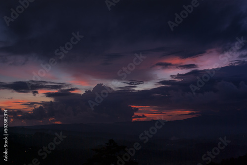 Colorful dark sunset landscape with bright orange, pink, yellow, purple, violet clouds on blue sky and silhouette dark hills in haze in Bali island, Munduk village.