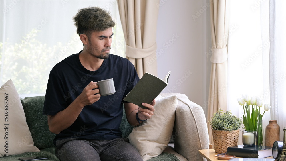 Peaceful millennial man drinking coffee and reading book on couch at home.