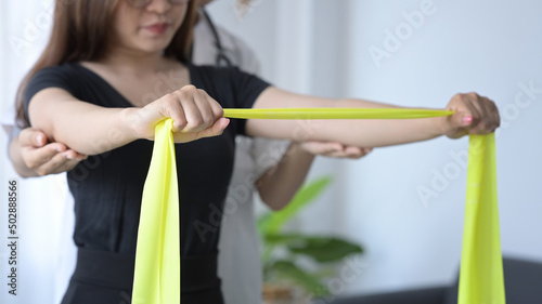 Female patient stretching yellow elastic band expander for rehabilitation exercises under physiotherapist supervision.