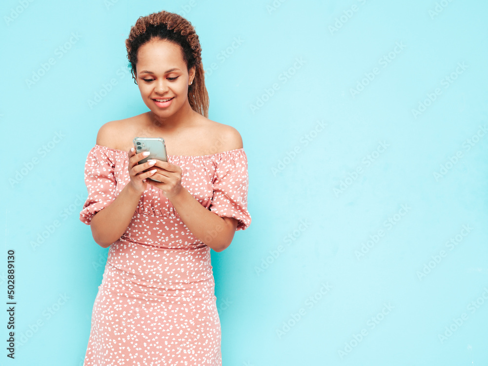 Beautiful black woman with afro curls hairstyle.Smiling model in trendy summer clothes. Sexy carefree female posing near blue wall in studio.Using smartphone apps.Looking at cellphone screen