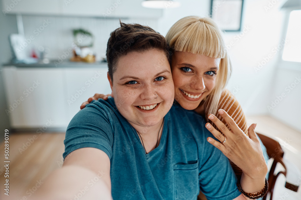 Happy lesbian showing off her engagement ring while taking selfie with her girlfriend at home.