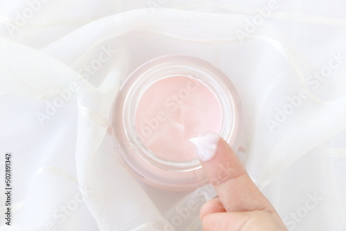 Top view of close up woman finger applying pink cream in jar on white fabric background