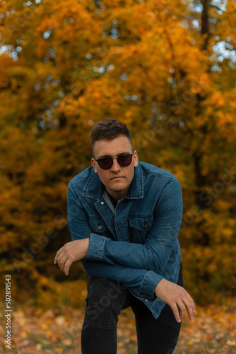 Handsome countryside man hipster with vintage sunglasses in jeans clothes stands and poses in autumn park with golden foliage © alones