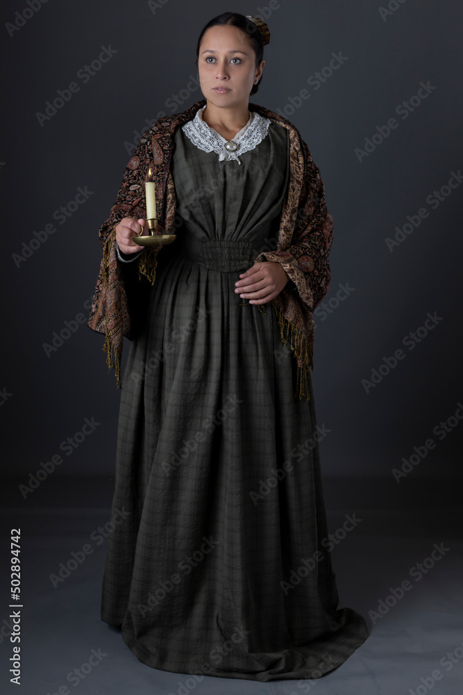 A Victorian working class woman standing alone holding a candle against a grey studio backdrop