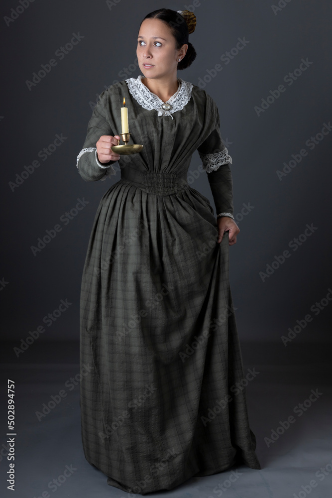 A Victorian working class woman standing alone holding a candle against a grey studio backdrop