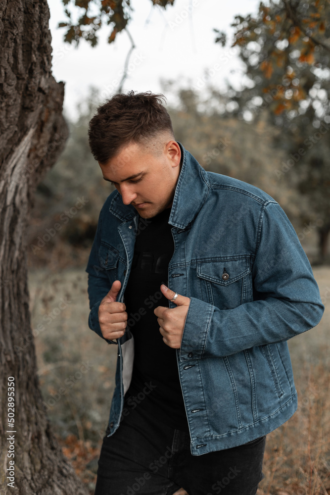 Handsome fashion model man with hairstyle in denim jacket with black t-shirt and jeans walks in the forest Handsome fashion model man with hairstyle in denim jacket with black t-shirt and jeans walks in the forest