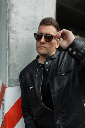 Cool stylish guy with hair in fancy black clothes with a leather jacket wears fancy glasses near a concrete pole in the city. Urban male portrait