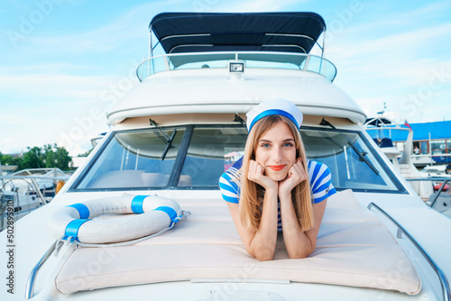 Attractive blond female skipper sits on deck with lifebuoy in striped dress in a sailor's cap on a sunny summer day on calm blue sea water. Luxurious summer adventure, active marine vacations.