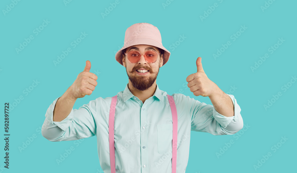 Cheerful funny charismatic young man showing thumbs up isolated on turquoise background. Bearded hipster in shirt, suspenders, panama and sunglasses looking at camera with funny expression on his face