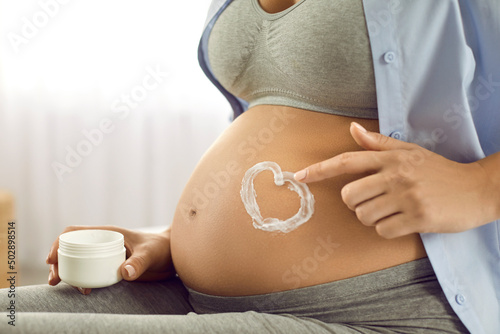 Pregnant woman on her naked tummy makes heart using moisturizer for stretch marks. Close up of belly of woman holding container with cream. Pregnancy, motherhood, preparation and expectation concept.