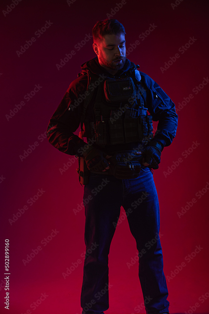 airsoft player in full gear with guns GG RK74. a man in headphones, body armor, with a backpack and a belt. red background. colored, blue-red light