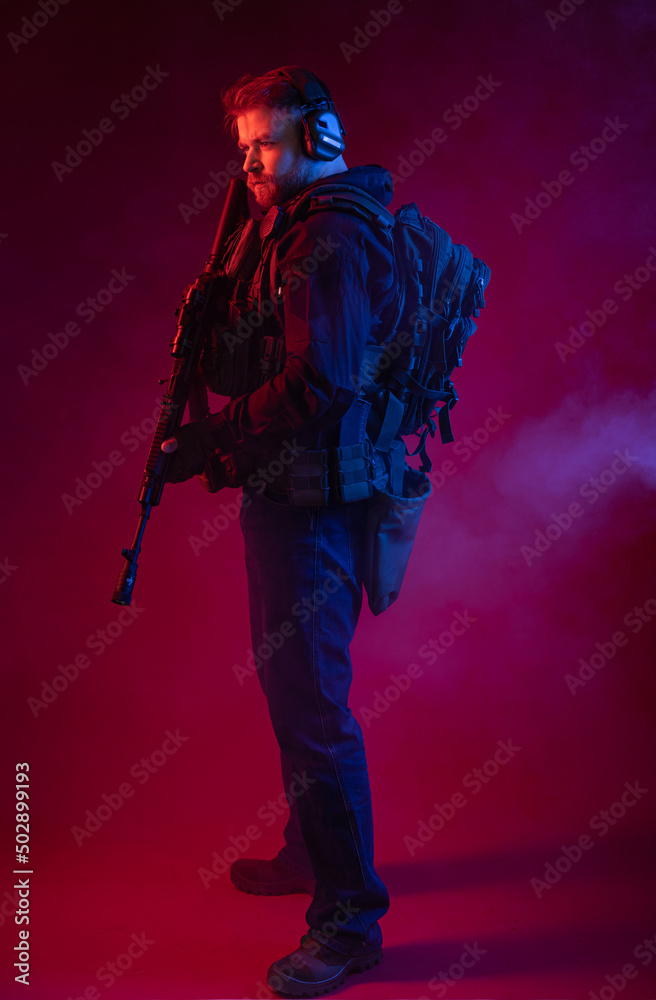 Obraz premium soldier in full gear with weapons. a man in headphones, body armor, with a backpack and a belt. red background. colored, blue-red light