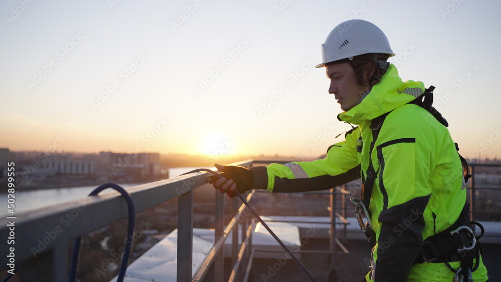 Industrial climber removes safety equipment from roof fence at sunset. Man in green overall finishes work on rooftop of skyscraper in evening