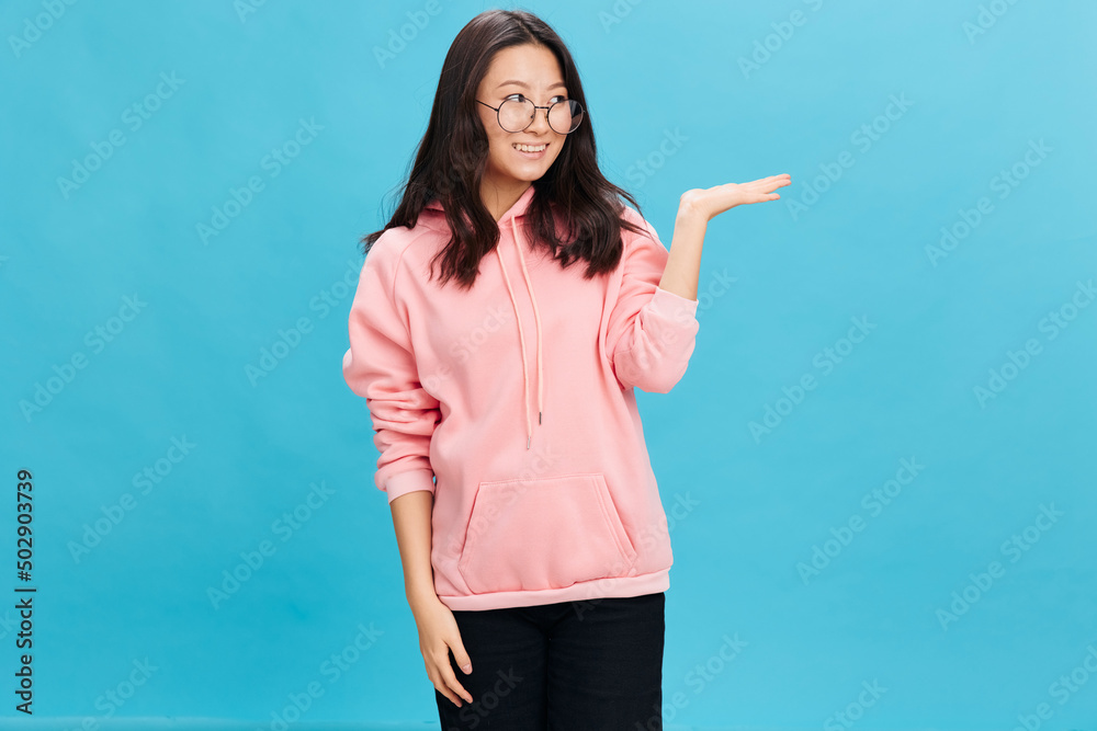 Joyful Asian young lady in pink hoodie sweatshirt wear round glasses holds invisible object on hand posing isolated on blue background. The best offer for ad. Eyewear for vision correction concept Joyful Asian young lady in pink hoodie sweatshirt wear round glasses holds invisible object on hand posing isolated on blue background. The best offer for ad. Eyewear for vision correction concept