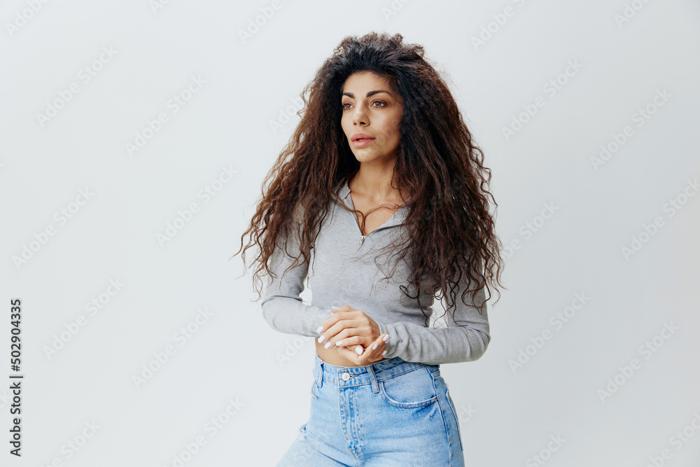 Sexy pretty curly Latin female wearing blue jeans gray sweatshirt folding hands looking aside. Copy space for clothing fashion brands, free place for ad. Studio shoot isolated white gray background Sexy pretty curly Latin female wearing blue jeans gray sweatshirt folding hands looking aside. Copy space for clothing fashion brands, free place for ad. Studio shoot isolated white gray background