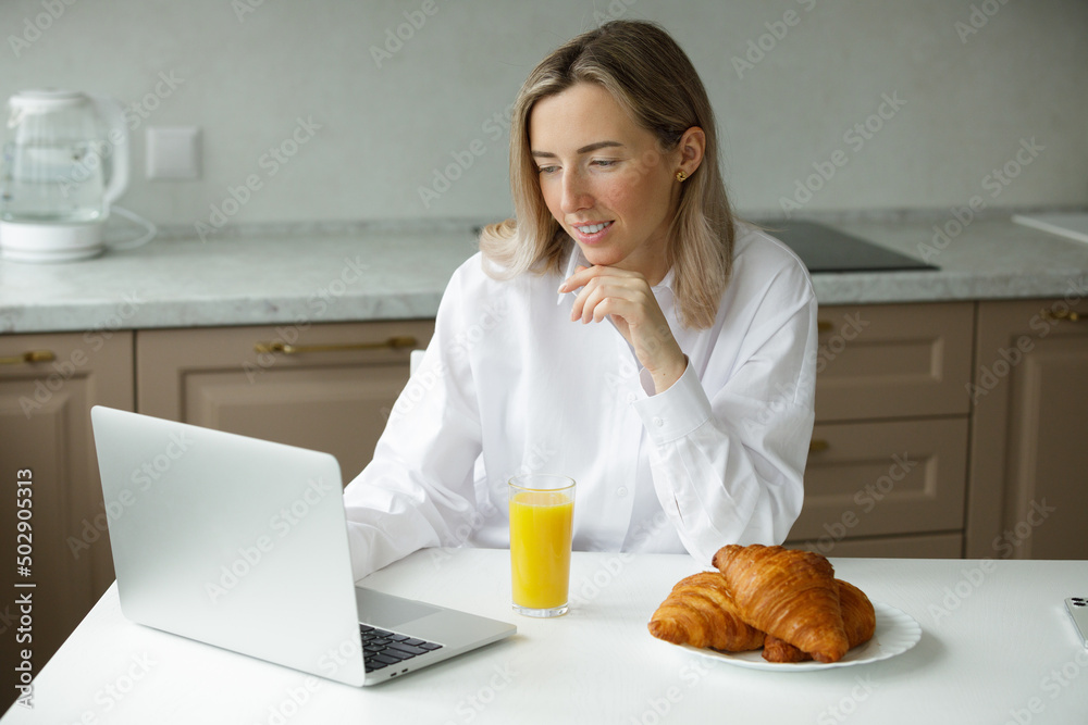 Young smiling girl in a white shirt eats croissants, drinks orange juice and works at a laptop