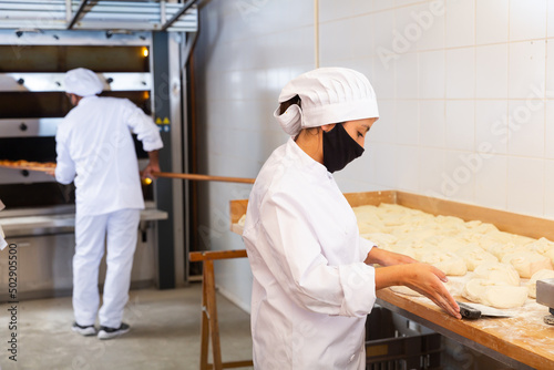 Female bakery worker wearing protective mask cuts dough with knife into identical loaves in bakery kitchen © JackF