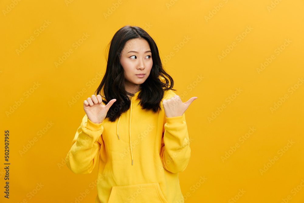 Pensive doubting Asian student young woman in yellow hoodie sweatshirt show at empty copy space for ad posing isolated on over orange studio background. Cool offer. People Emotions concept Pensive doubting Asian student young woman in yellow hoodie sweatshirt show at empty copy space for ad posing isolated on over orange studio background. Cool offer. People Emotions concept