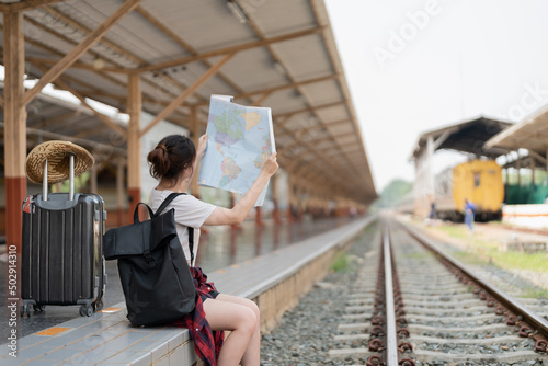 Young asian woman using generic local map, siting alone at train station platform with luggage. Summer holiday traveling or young tourist backpack traveler concept