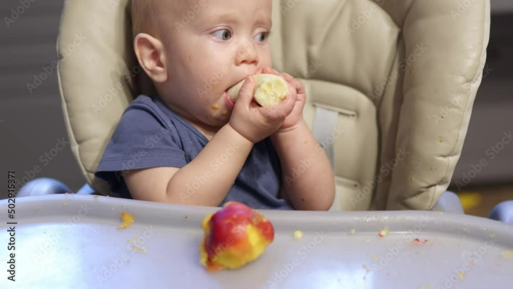 portrait eating baby fruit at home.healthy food, lunch time. kid child biting peach, holding it in hand. baby making funny face tasting sweet fruit, eating pieces, sitting at feeding chair table. portrait eating baby fruit at home.healthy food, lunch time. kid child biting peach, holding it in hand. baby making funny face tasting sweet fruit, eating pieces, sitting at feeding chair table.