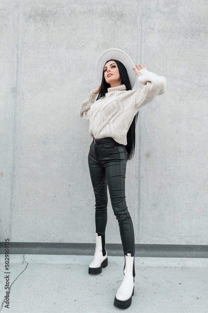 Fashionable beautiful woman model in stylish clothes with knit sweater, leather pants and shoes stands near the gray wall on the street and adjusts her hat Fashionable beautiful woman model in stylish clothes with knit sweater, leather pants and shoes stands near the gray wall on the street and adjusts her hat