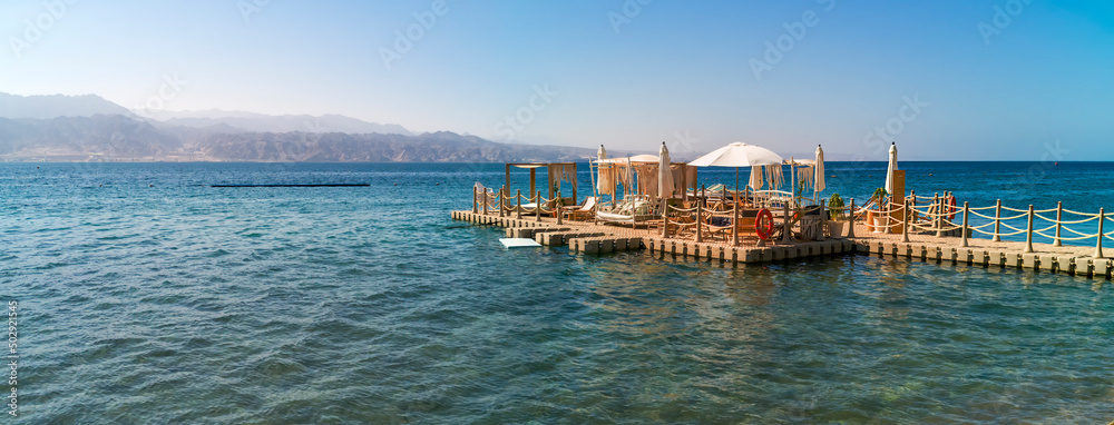 Fototapeta premium Resting area with artificial floating island and tables and chairs on relaxing public beach of the Red Sea, Middle East. Concept of bliss and happy vacation