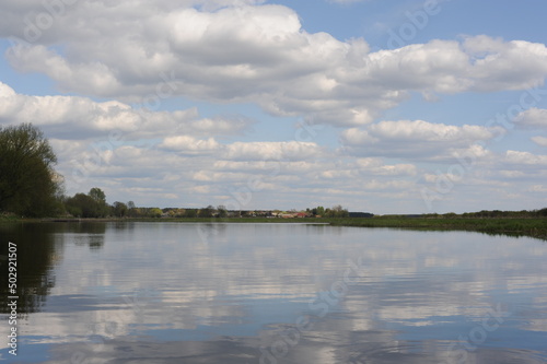 Picturesque and peaceful cloudscape with white clouds and blue sky reflecting in the river in spring or summer in Poland © piotrmilewski
