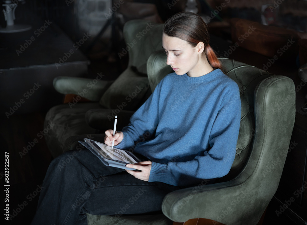 Girl sits in the lobby of a hotel or restaurant and draws or works on electronic tablet.
The concept of inspiration, creativity, self-development, hobby, modern art. Modern technologies. Remote work