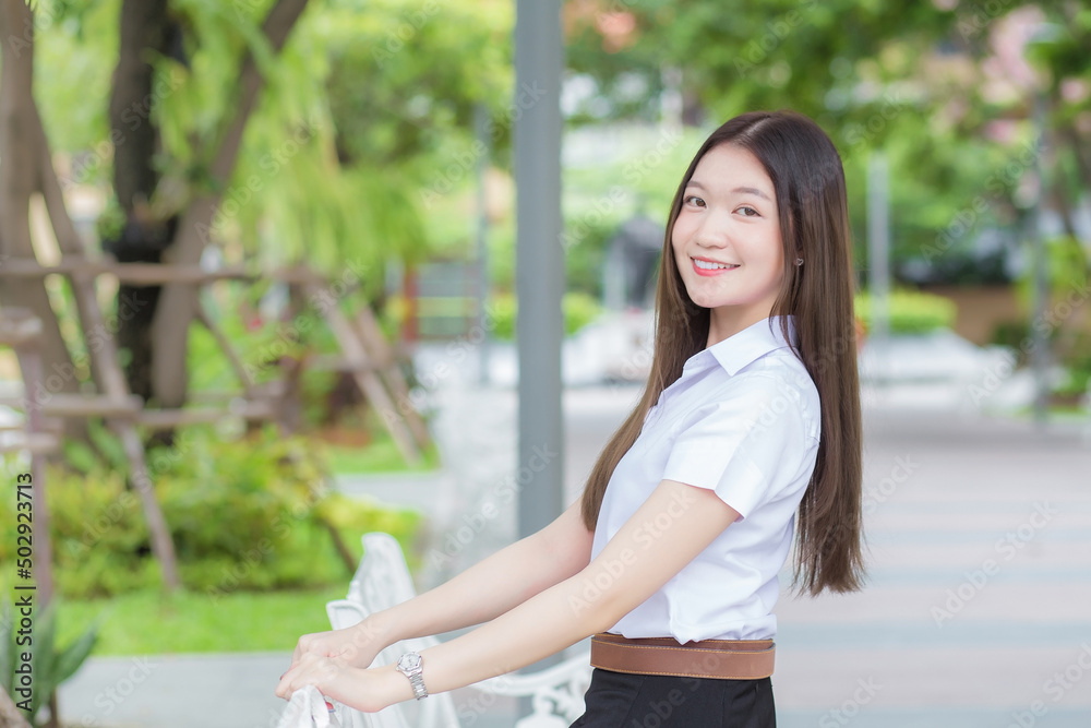 Portrait of an adult Thai student in university student uniform. Asian beautiful girl sitting smiling happily at university with a background of garden trees. Portrait of an adult Thai student in university student uniform. Asian beautiful girl sitting smiling happily at university with a background of garden trees.