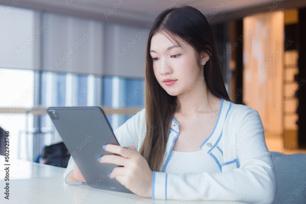 Beautiful young age Asian professional woman with long hair in white shirt holds tablet her smiling happily while she sits on chair in workplace office with glassed building as a background. Beautiful young age Asian professional woman with long hair in white shirt holds tablet her smiling happily while she sits on chair in workplace office with glassed building as a background.