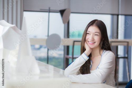 Asian young woman with long hair smiles while looking at the camera while she sits on chair in workplace with glassed building as a background.