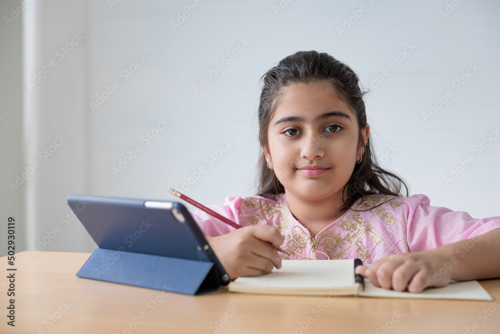 portrait cute Indian girl writing on notebook and online learning class from tablet, education concept portrait cute Indian girl writing on notebook and online learning class from tablet, education concept