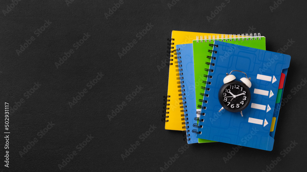 Obraz premium A stack of colored school notebooks and an alarm clock on a school chalkboard background. Back to school concept, banner