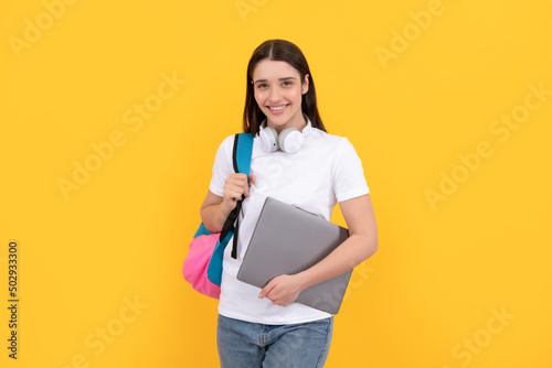 young girl with backpack hold laptop in headphones on yellow background, education