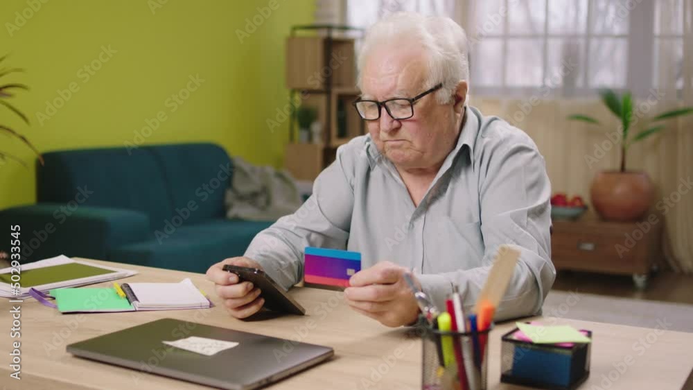 Charismatic old man at home using the smartphone to pay something online from the credit card he holding his credit card and type the card number Charismatic old man at home using the smartphone to pay something online from the credit card he holding his credit card and type the card number