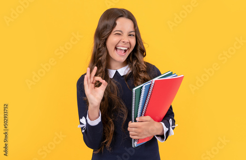 happy winking kid hold school copybooks studying on yellow background show ok gesture, school.