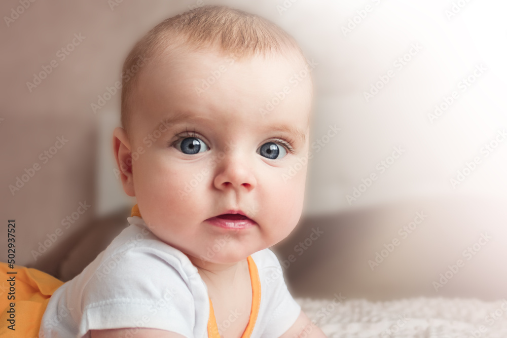 Portrait of a baby girl with big blue eyes lying on her stomach on the bed and looking at the camera