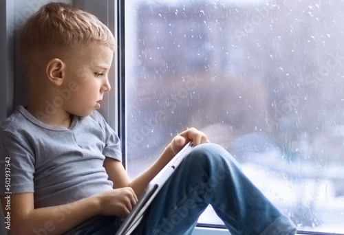 a little fair-haired boy in a gray T-shirt and jeans sits on the windowsill, holds a tablet in his hands and looks sadly out the window, it's raining. Social concept. Stay home. photo