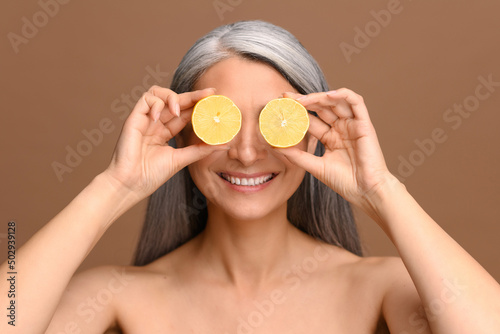 Fun portrait of middle-aged gray-haired half-naked Asian female covering eyes with halves of lemon standing isolated on dark-beige, well-looking lady using citrus acid for skin care