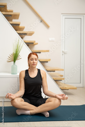 Young healthy beautiful woman in sportive clothes practicing yoga sitting on yoga mat meditating at home.