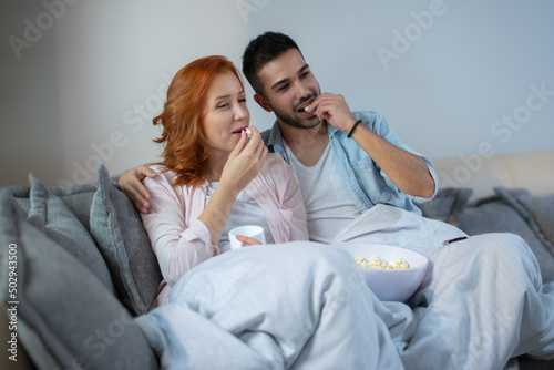 Happy couple eating popcorn and watching a movie on TV while lying on a comfortable sofa at home.