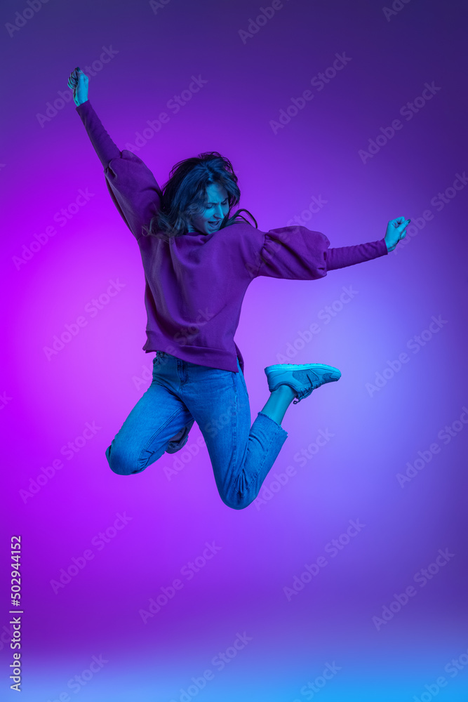 Full-length portrait of young excited girl, student jumping isolated over purple background in neon. Concept of emotions, facial expression, art, beauty