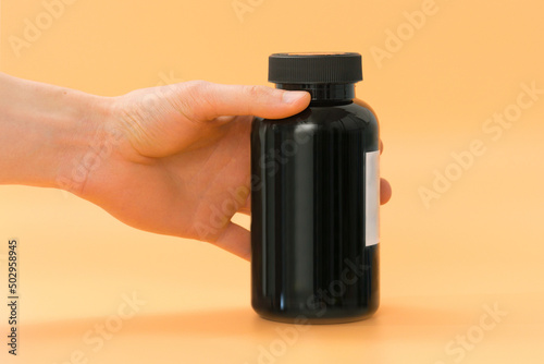 A tall black plastic supplement bottle is in the man's hand on a yellow background. Workout. Vitamin. Pill. Pack. Prescription. Box. Healthy. Standing. Template