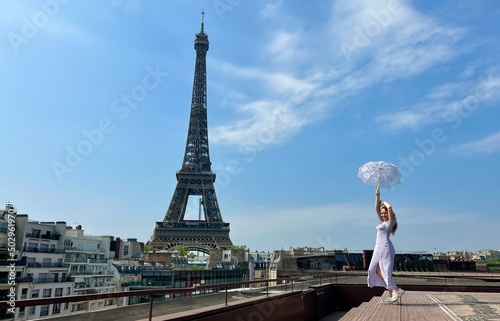 girl in a blue dress stands on the background of the Eiffel Tower a lot of space for text she is happy in hands holding a sun umbrella raising her hands Up looks like Mary Poppins. High quality photo
