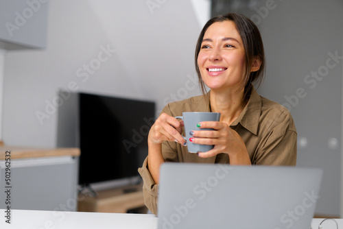 Smiling young cheerful asian woman enjoying morning coffee while looking away while working at the laptop, talking with her virtual friend, sitting at the desk in home office
