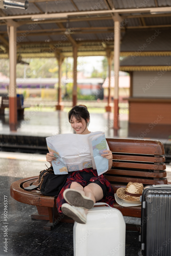 Fototapeta premium Happy young asian woman traveler or backpacker using map choose where to travel with luggage at train station, summer vacation travel concept